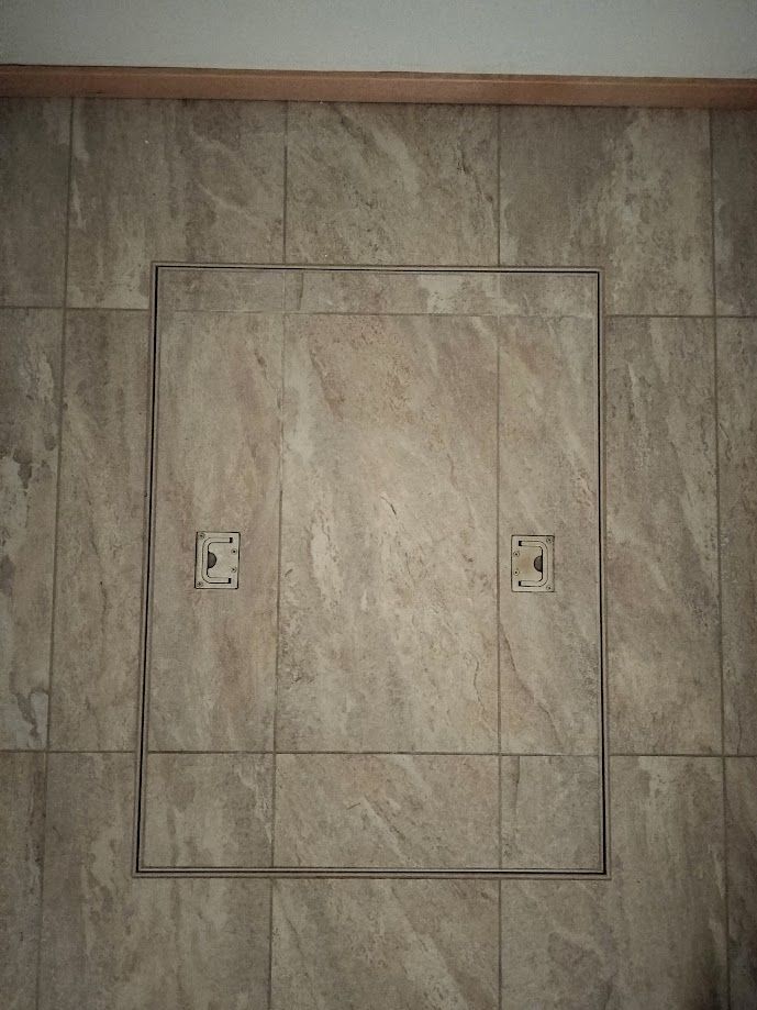 A rectangular access panel with matching tiled front, centered on a tiled wall with two small latch mechanisms visible.