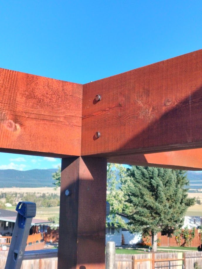 A close-up of a corner joint on a reddish-brown wooden pergola structure with metal bolts securing the beams to the post.