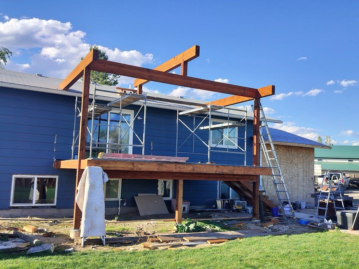 A blue house under construction, featuring a partially built elevated wooden deck and roof frame with scaffolding nearby.
