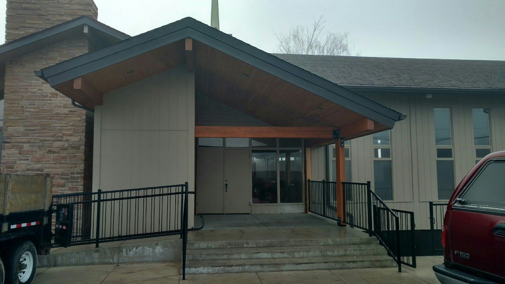 A building entrance featuring a gable roof, wood-toned supports, and a black metal railing leading to a set of stairs.