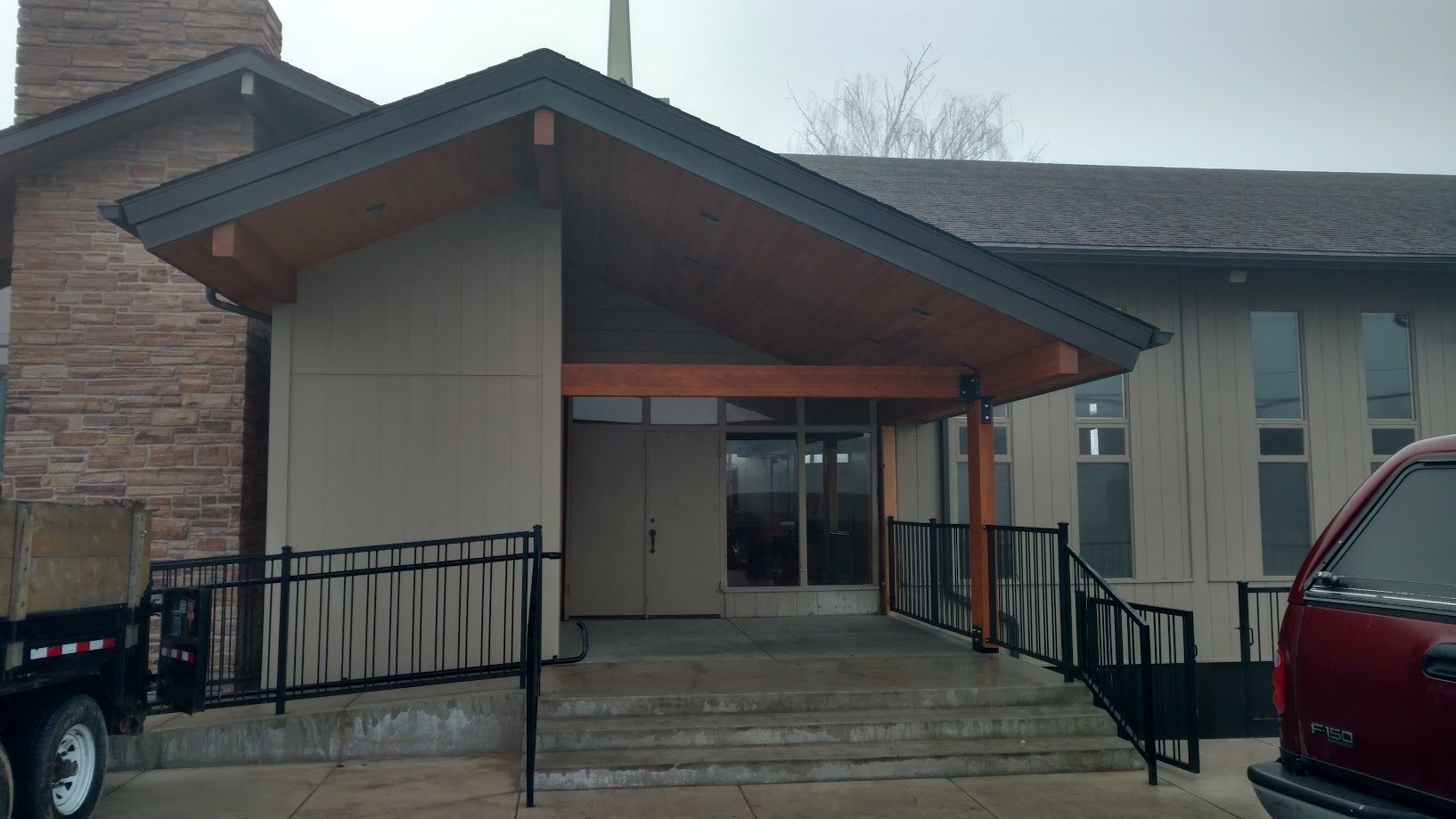 The entrance to a beige building with a dark shingled roof, a wooden-beam porch, and a black metal railing ramp.