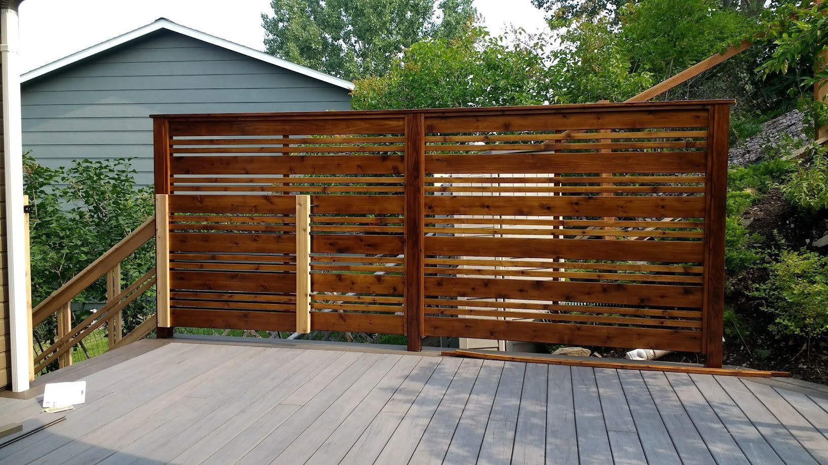 A horizontal wood privacy screen stands on a grey deck, with two unpainted support posts visible on the left side.