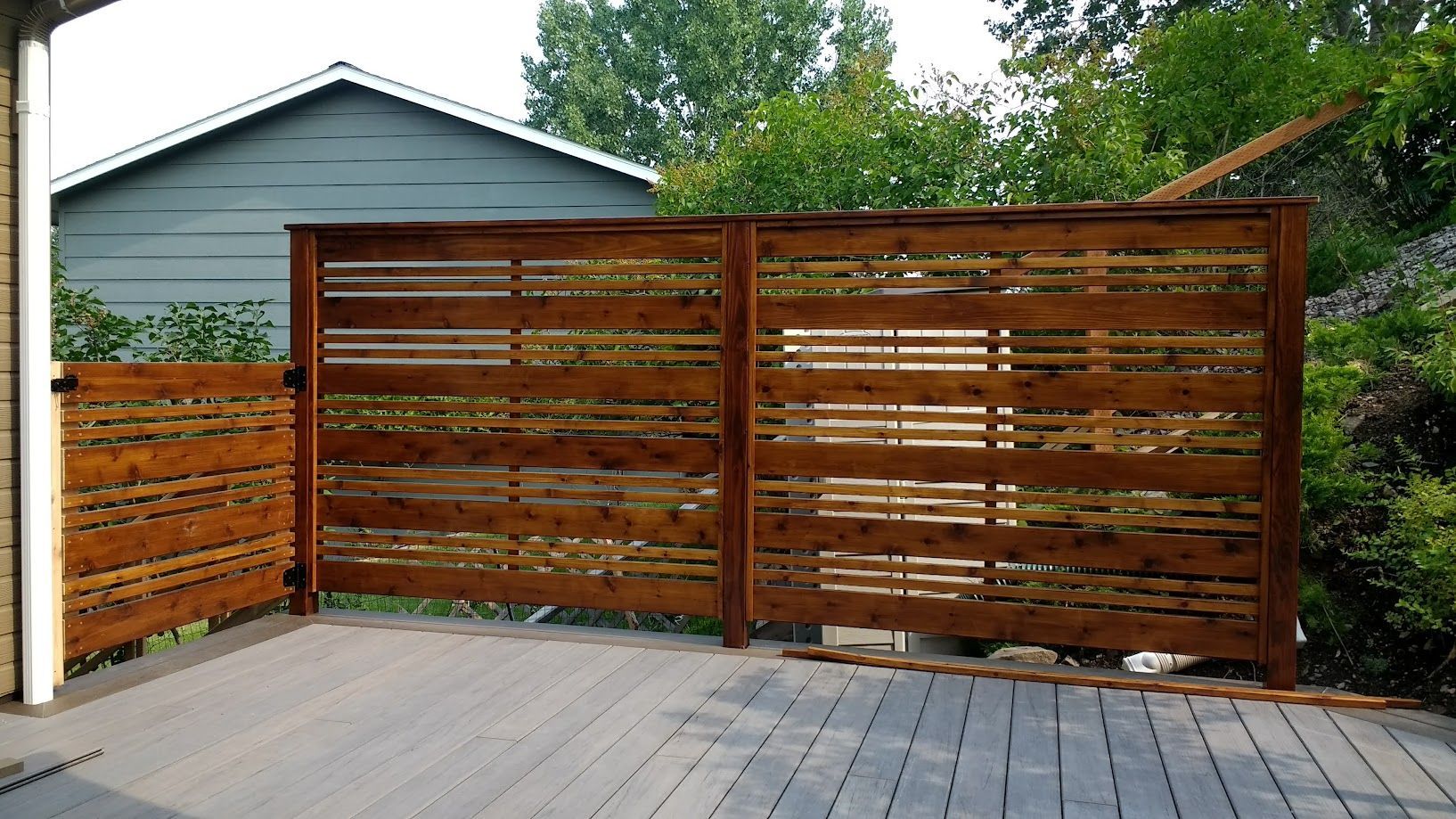 A horizontal-slat wooden privacy fence on a grey composite deck, situated in front of a blue house and green foliage.