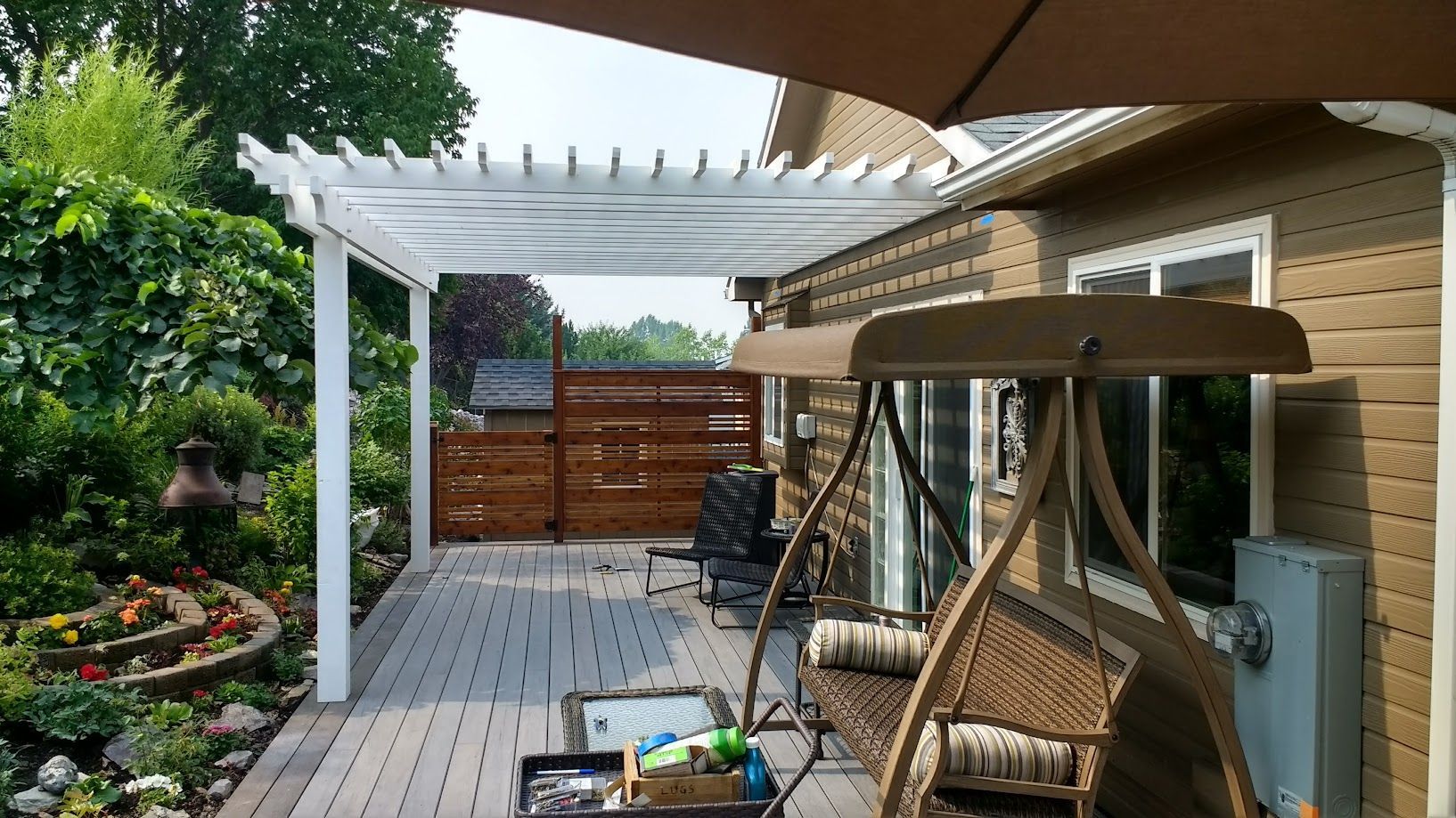 A patio with a white pergola, a hanging outdoor swing, and a wooden privacy screen next to a house with beige siding.
