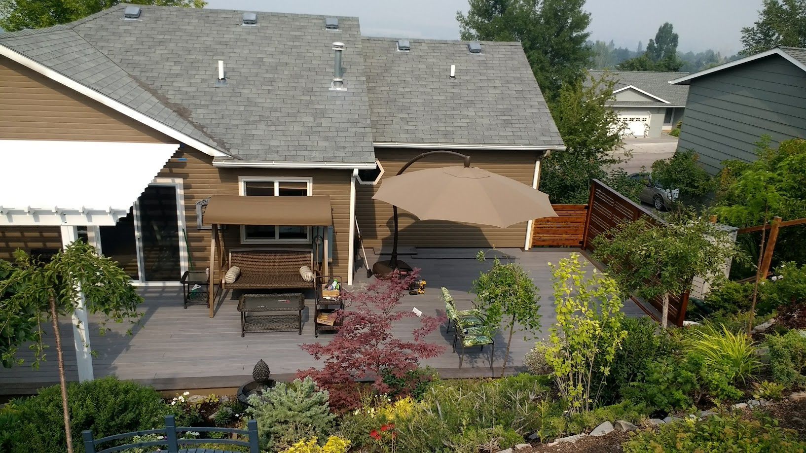 A backyard patio with a wooden swing, a brown umbrella, and landscaping, viewed from an elevated angle.