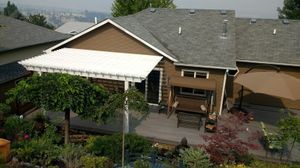 A high-angle view of a backyard patio with a white pergola, a porch swing, a large tan umbrella, and lush landscaping.
