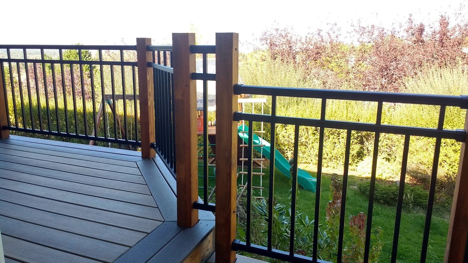 A wooden deck with black metal railings overlooking a backyard with a green children's slide.