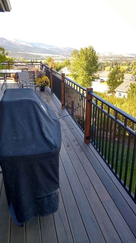 A covered grill sits on a wooden deck with a black railing, overlooking a suburban neighborhood and distant mountains.