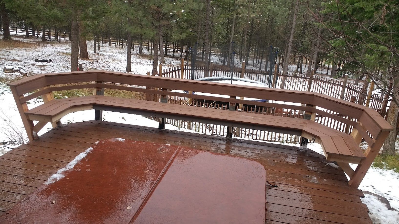 A wooden deck with a built-in curved bench and a hot tub cover, surrounded by a snowy forest.