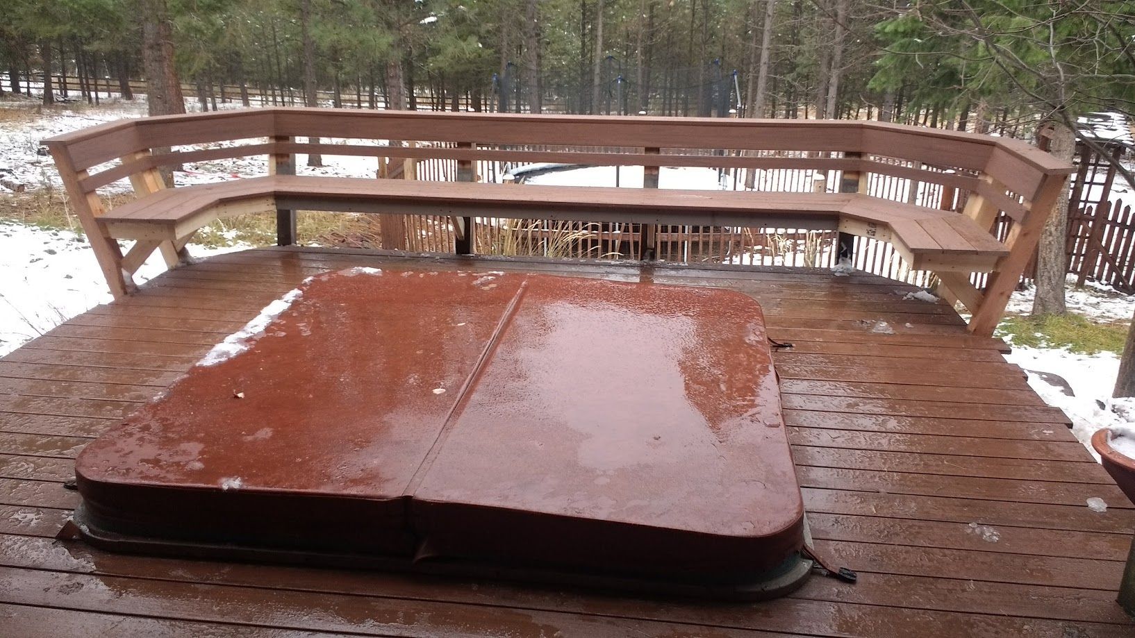 A reddish-brown hot tub cover on a wooden deck with a curved bench, surrounded by a snowy forest.