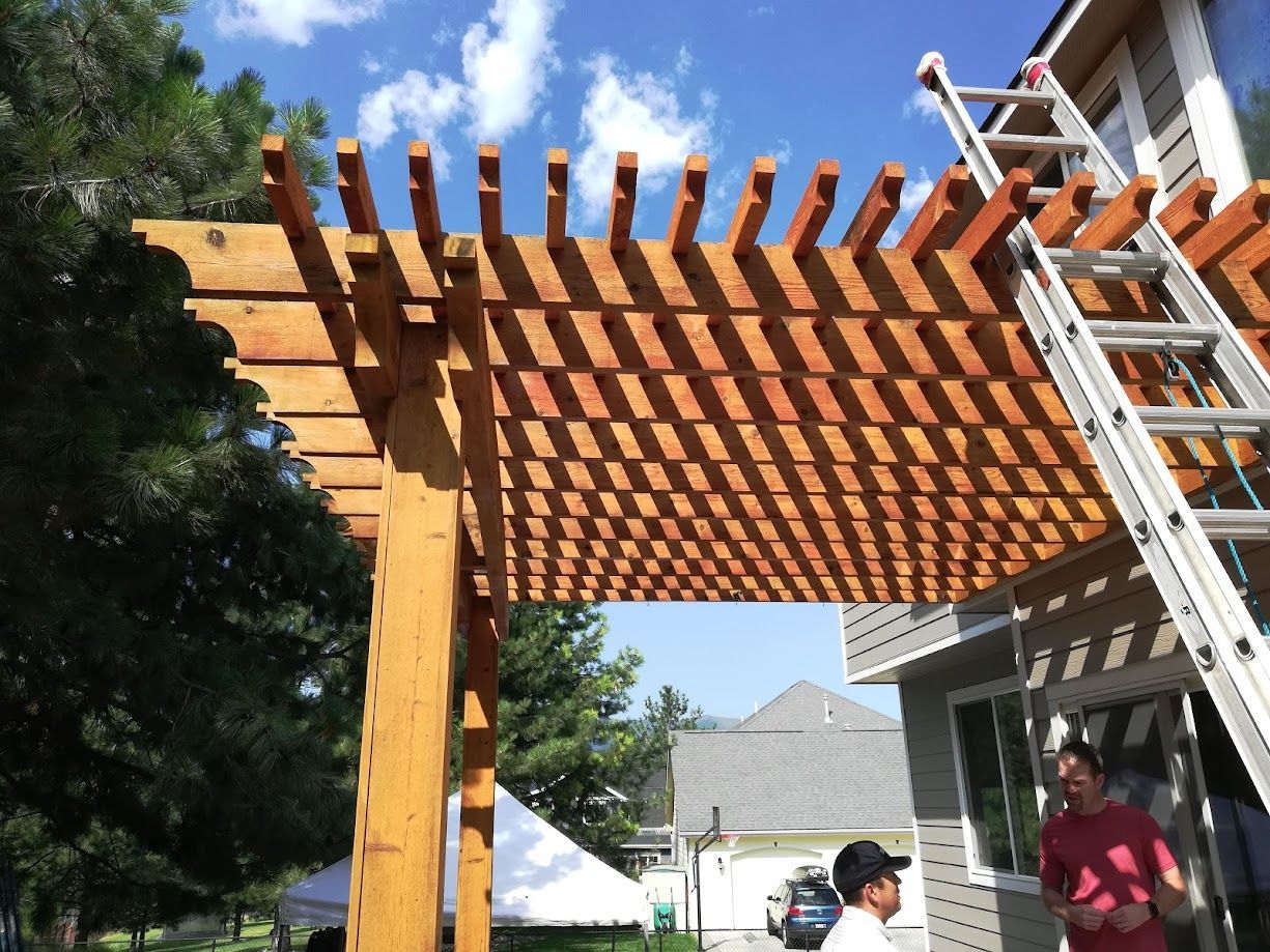 A wooden pergola attached to a house exterior, with a ladder leaning against the structure and two people visible nearby.