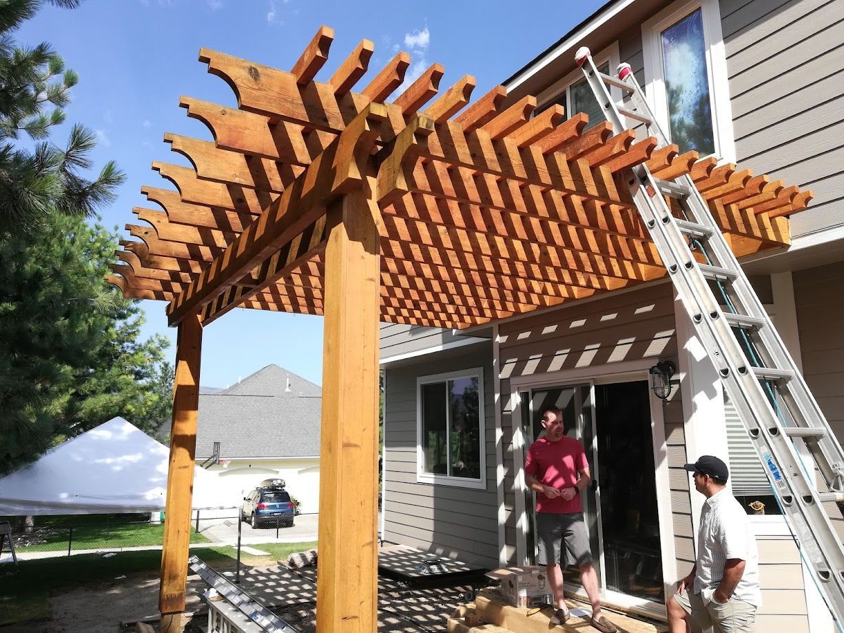 Two people stand beside a wooden pergola attached to a suburban house with a ladder leaning against the structure.
