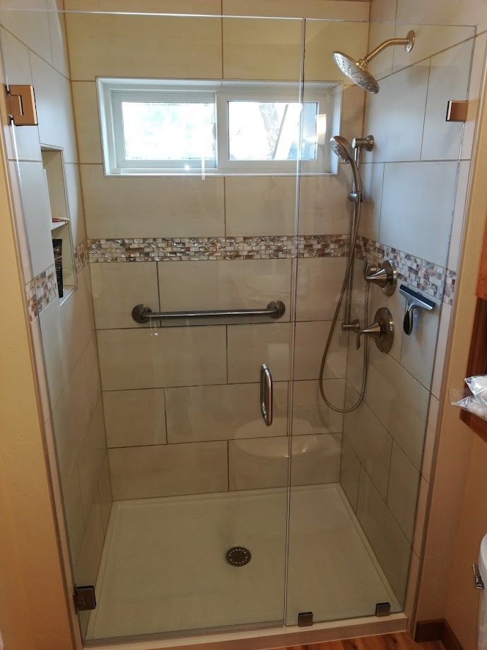A glass-enclosed walk-in shower with beige tile walls, a horizontal grab bar, a window, and a wall-mounted showerhead.