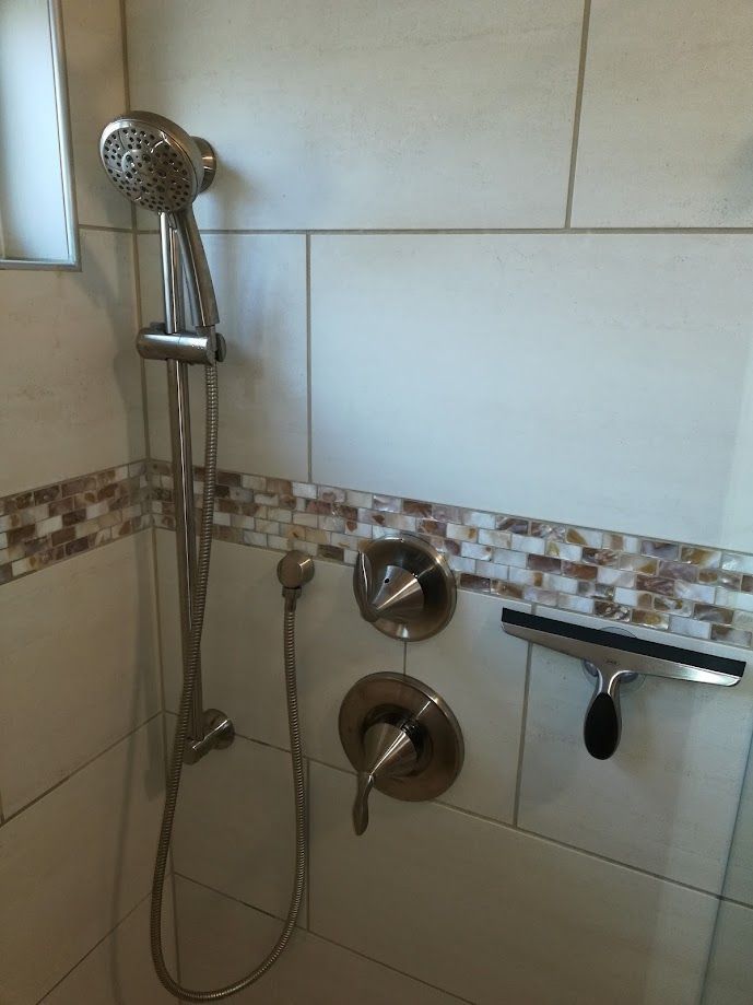 A modern shower interior featuring a handheld spray head, control valves, and a squeegee mounted on tiled walls.