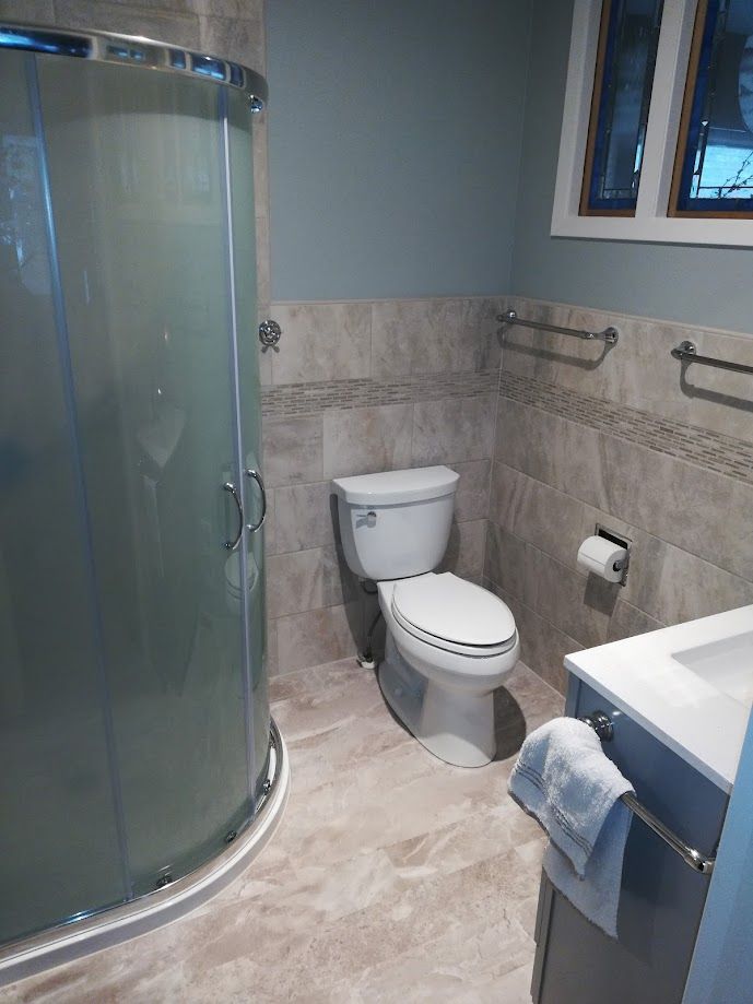 A renovated bathroom featuring a curved glass shower, a white toilet, and a vanity with tile flooring and walls.