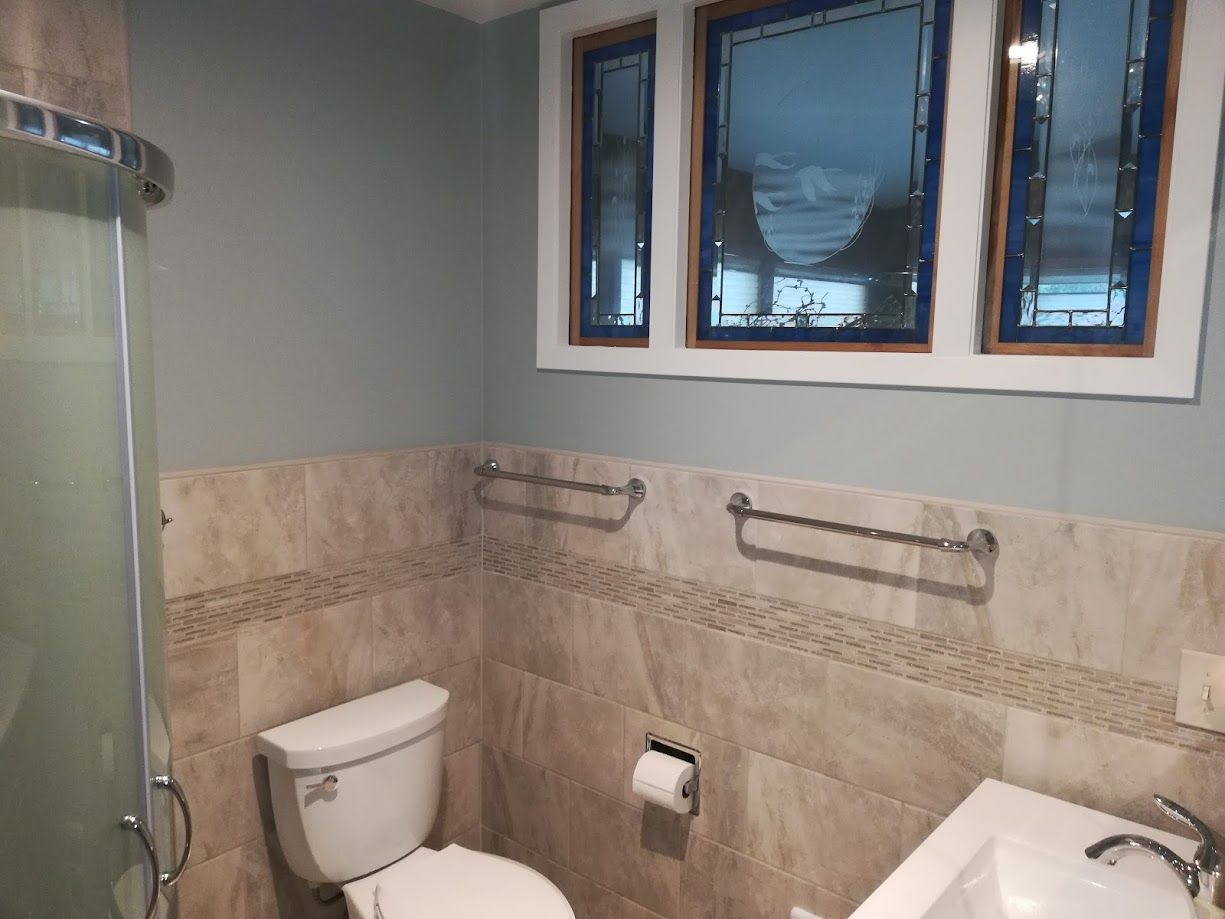 A bathroom with a corner shower, toilet, and sink, featuring beige tile wainscoting and three windows with blue frames.