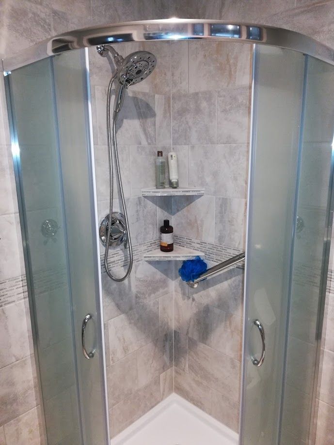 A curved glass walk-in shower with grey tiled walls, a handheld showerhead, and two corner shelves holding toiletries.