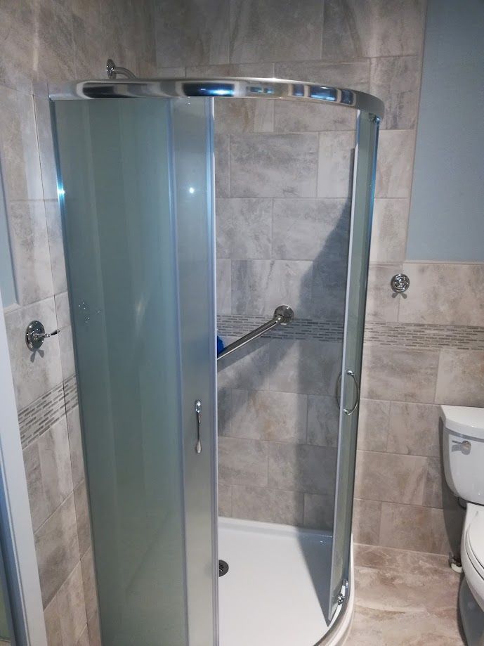 A corner shower stall with frosted glass doors and a chrome handle, set against grey tiled walls in a bathroom.