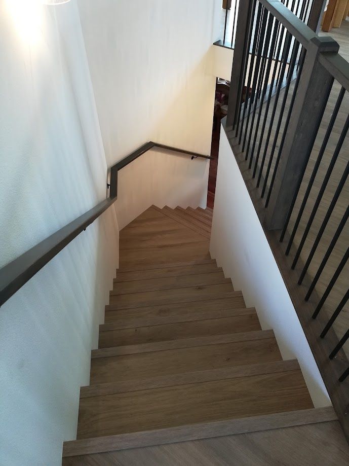 A downward view of a wooden staircase with dark railings, leading to a landing and a doorway in a brightly lit hallway.
