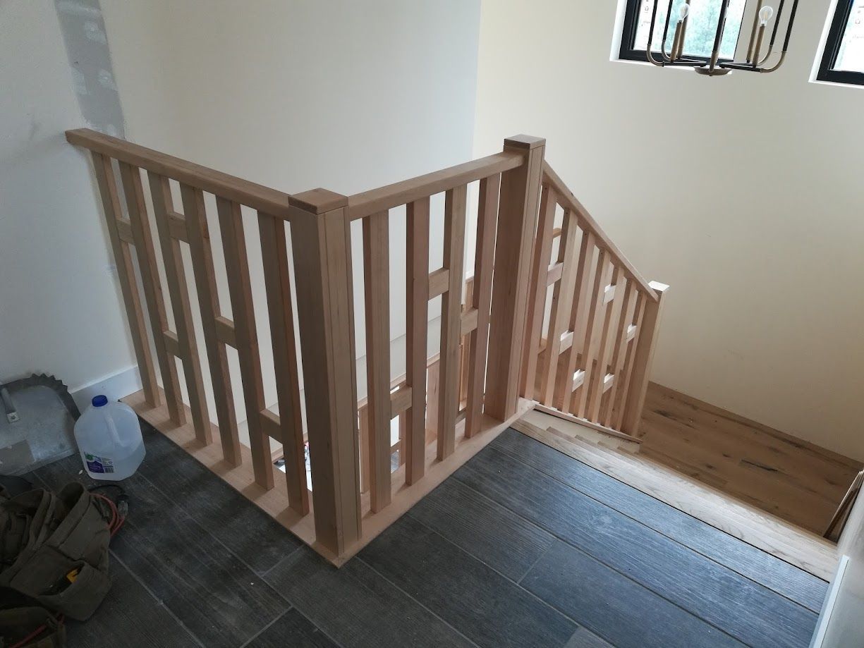 New light-wood staircase railing with vertical slats on a landing, overlooking stairs with dark wood and grey tile floors.