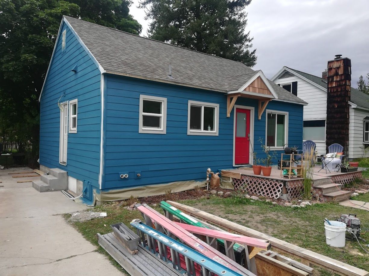 A blue-sided cottage house with a red front door, an unfinished wooden porch overhang, and construction equipment outside.