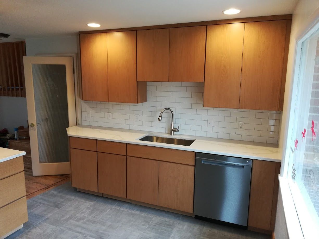 A kitchen featuring light wood cabinets, a white countertop, a stainless steel dishwasher, and a white tiled backsplash.