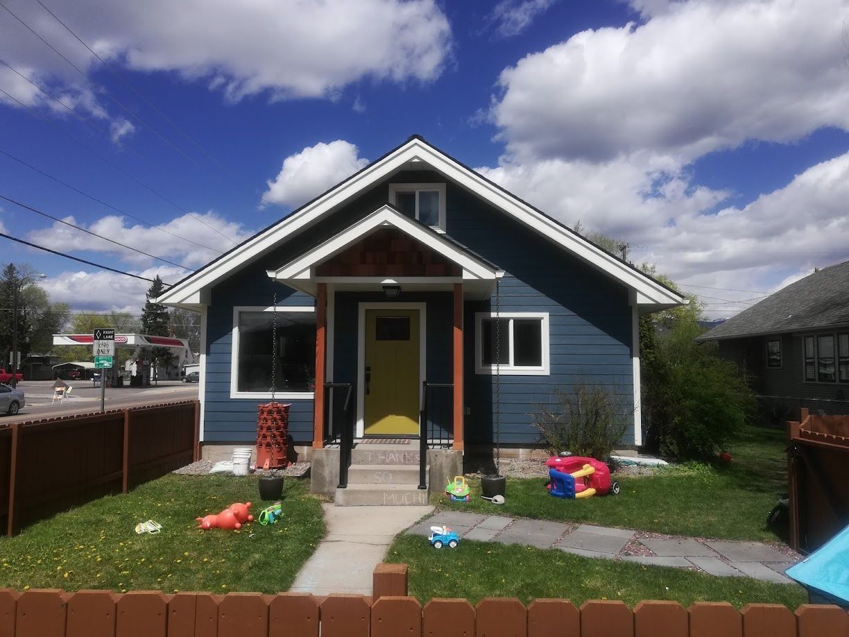 A blue, single-story house with a yellow front door, wooden porch supports, and toys scattered on the front lawn.