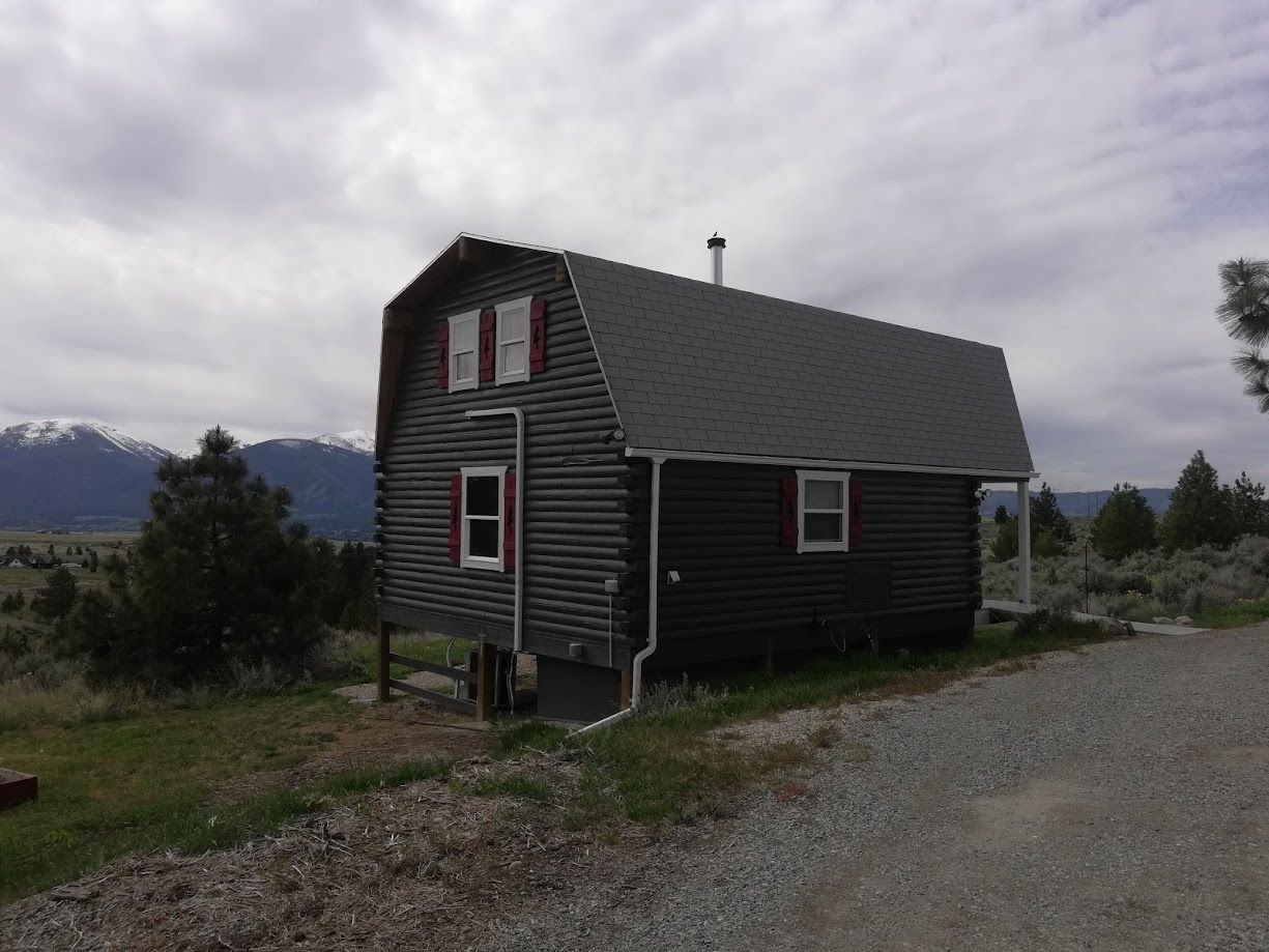 A dark-wood cabin with a gambrel roof and red-trimmed windows sits on a gravel lot before snow-capped mountains.