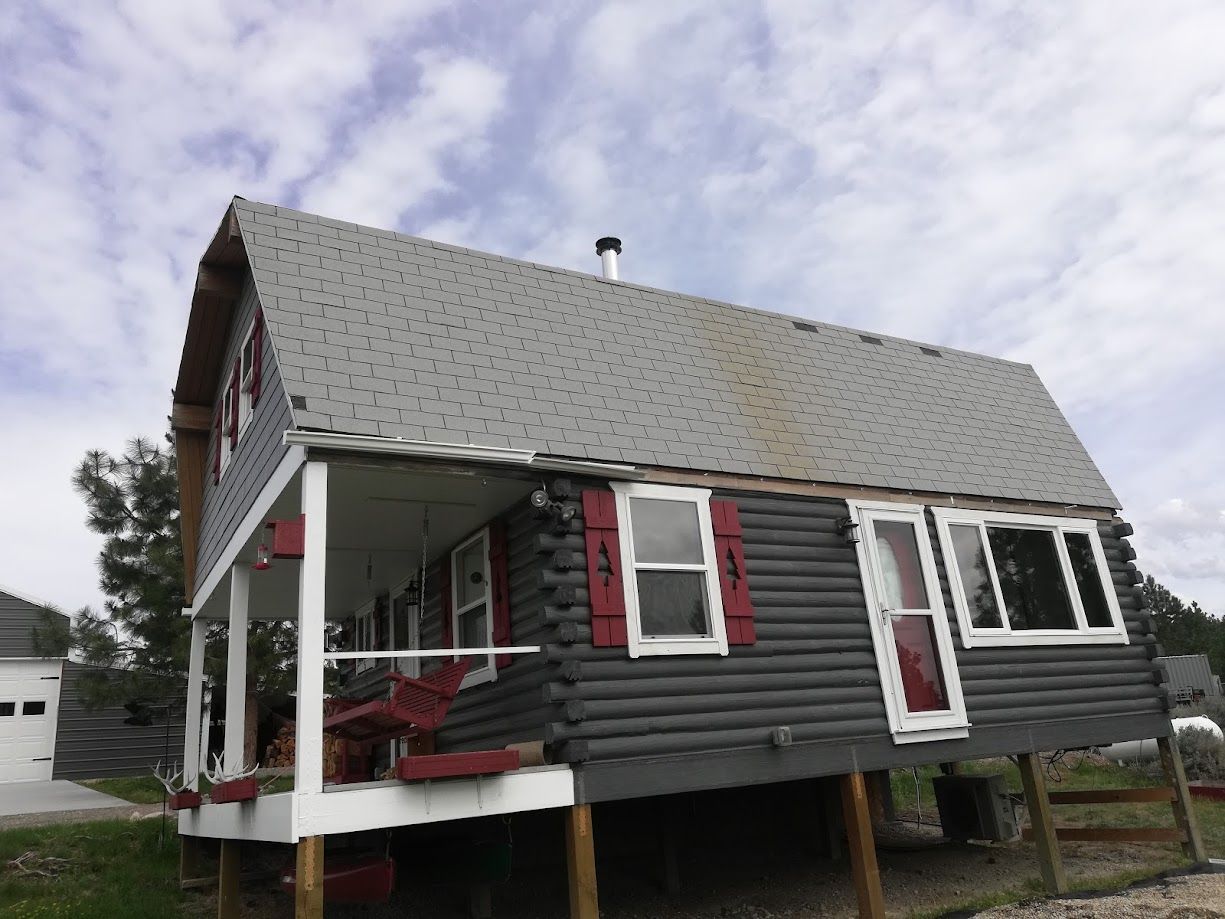 A rustic, dark-gray log cabin on raised pillars featuring a front porch with a red bench and matching red window shutters.