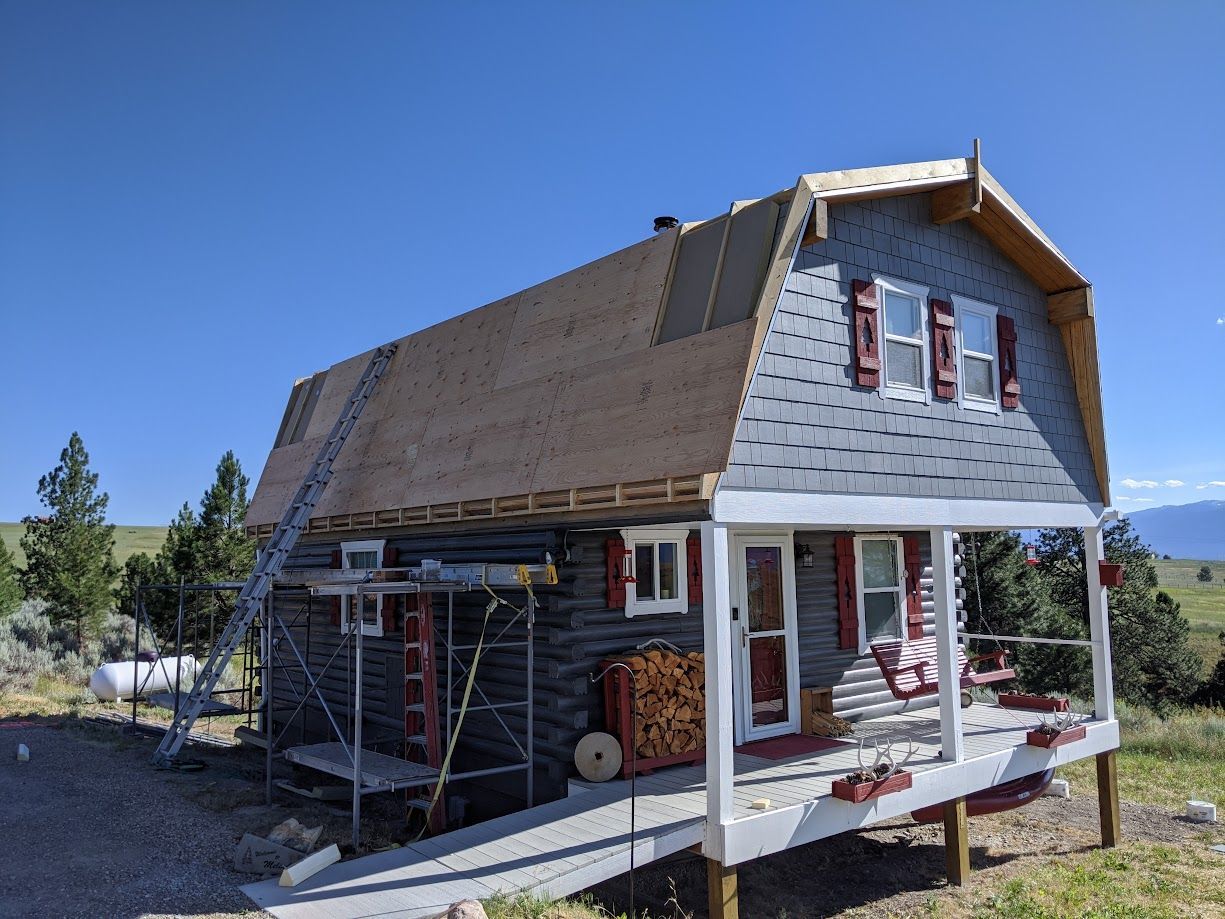 A rustic log cabin with a partial gambrel roof, under repair with visible underlayment and a ladder leaning against it.