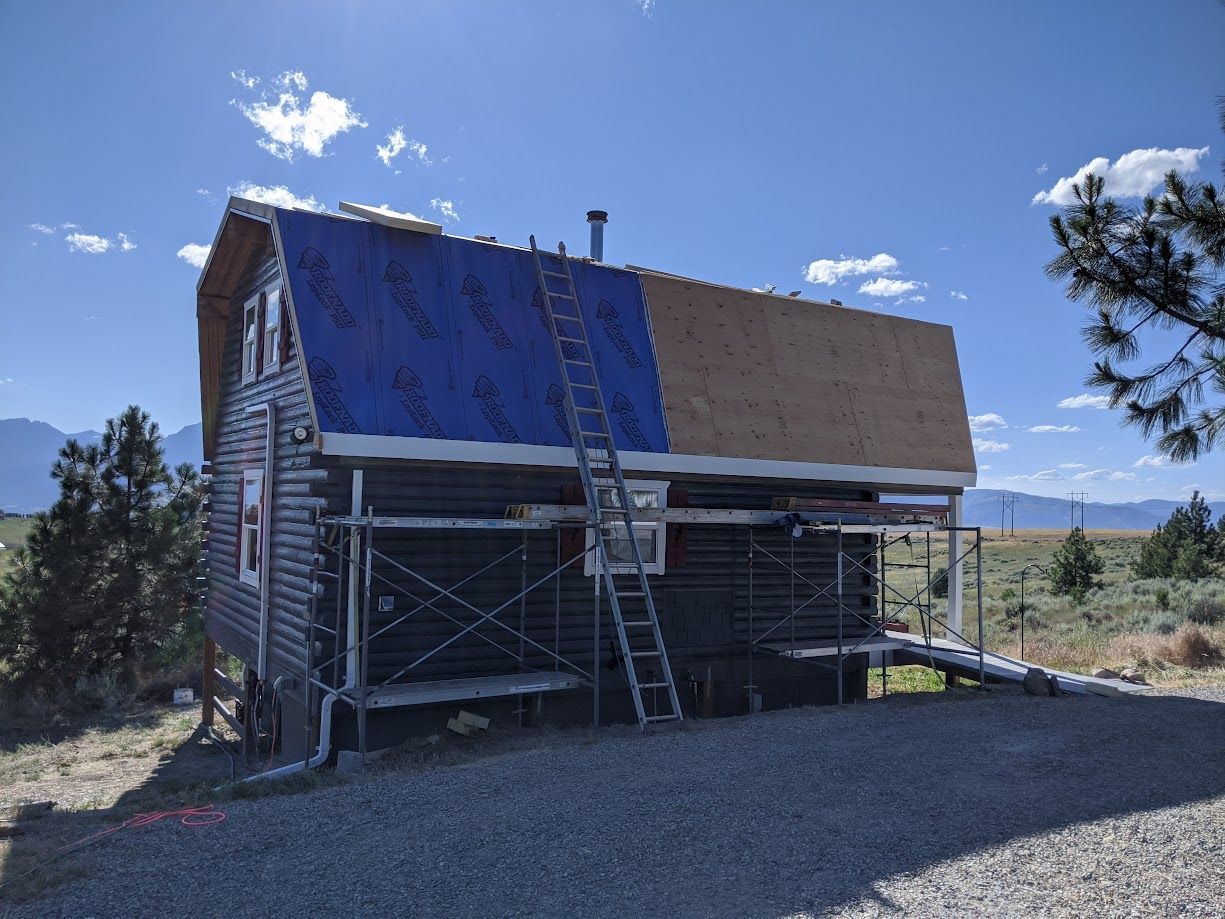 A partially re-roofed log cabin in a rural landscape, featuring blue underlayment on one side and bare plywood on the other.