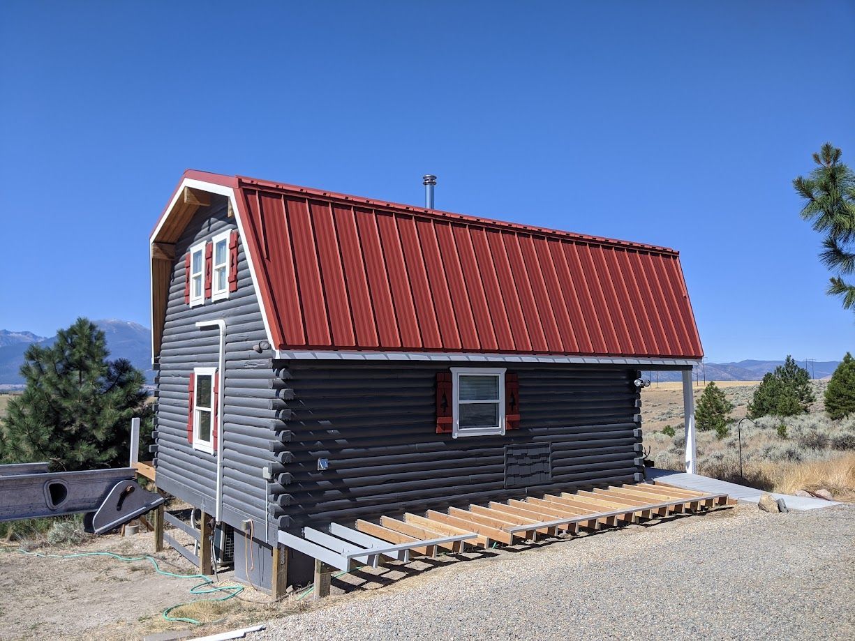 A small, gray log cabin with a red metal gambrel roof and an unfinished wooden deck in a dry, mountainous landscape.