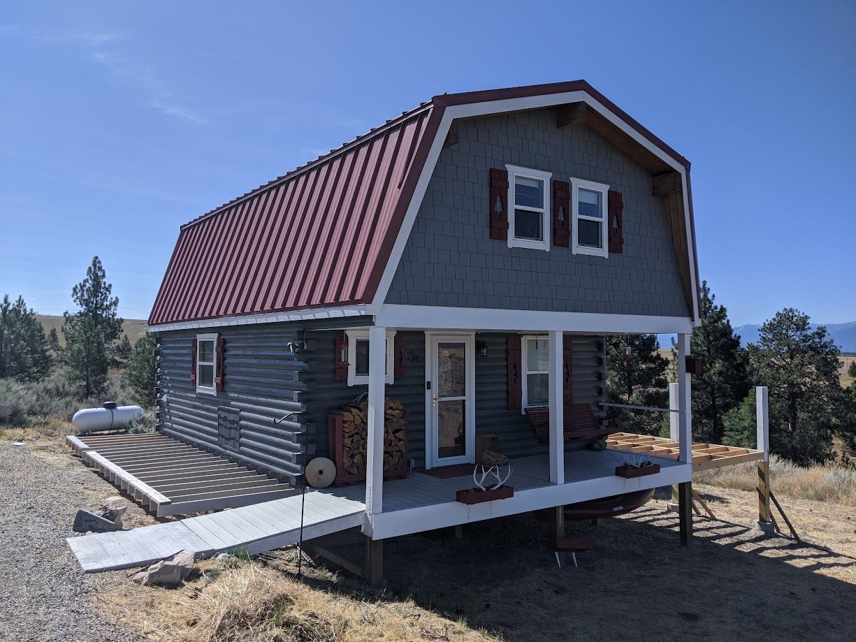 A small, grey log cabin with a red gambrel roof and front porch, set in a dry, grassy, treed landscape under a blue sky.
