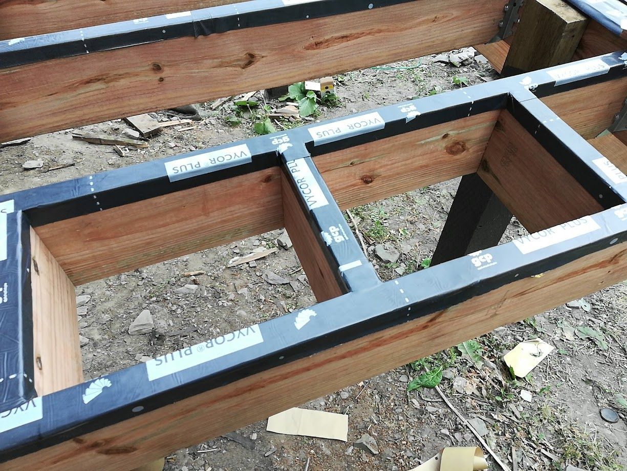 A wood deck frame under construction, with black joist tape applied to the top of the wooden beams.