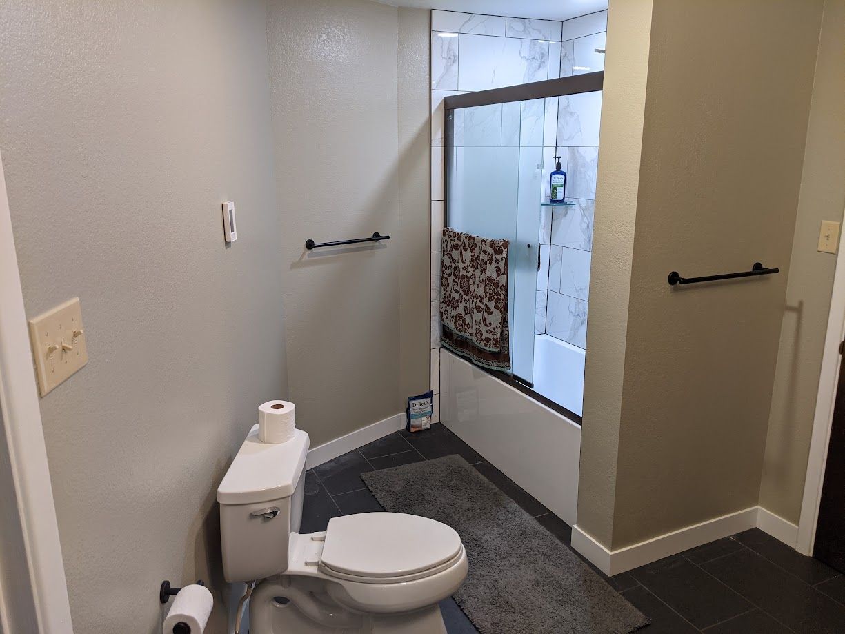 A bathroom featuring a toilet, a dark gray floor rug, and a walk-in shower with glass doors and a hanging patterned towel.