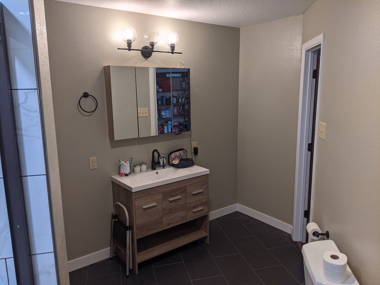 A bathroom vanity with a wood-grain cabinet, white sink, mirror cabinet, and vanity light against a taupe wall.