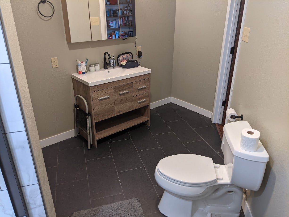 A bathroom with a wooden vanity, sink, wall mirror, and white toilet on a dark tiled floor.