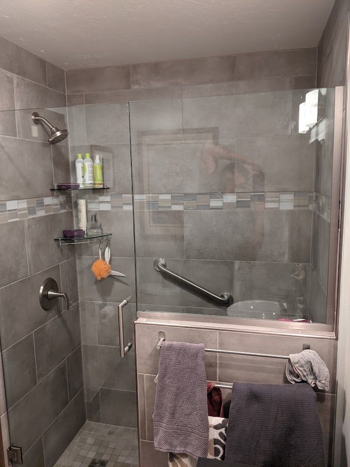 A walk-in shower with gray tile, a glass door, built-in shelving, a shower head, and an installed safety grab bar.
