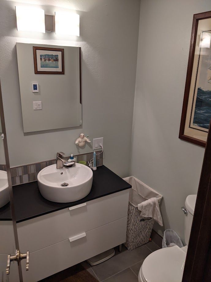 A bathroom featuring a white vanity with a black countertop, a vessel sink, a mirror, and a toilet next to a laundry hamper.