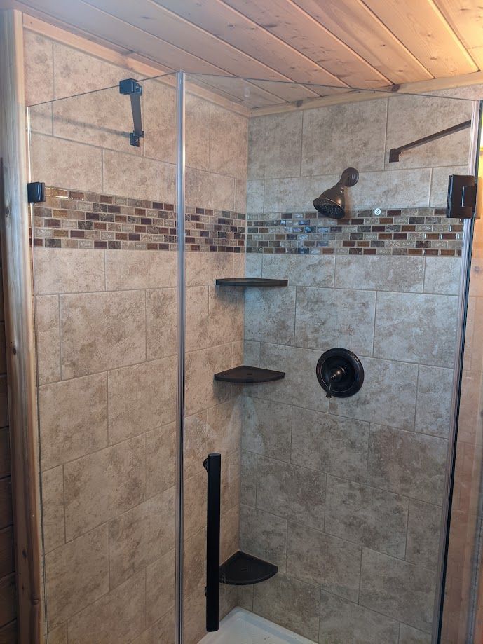 Modern corner shower with tan stone-look tiles, a mosaic accent strip, glass door, and black fixtures in a wooden room.