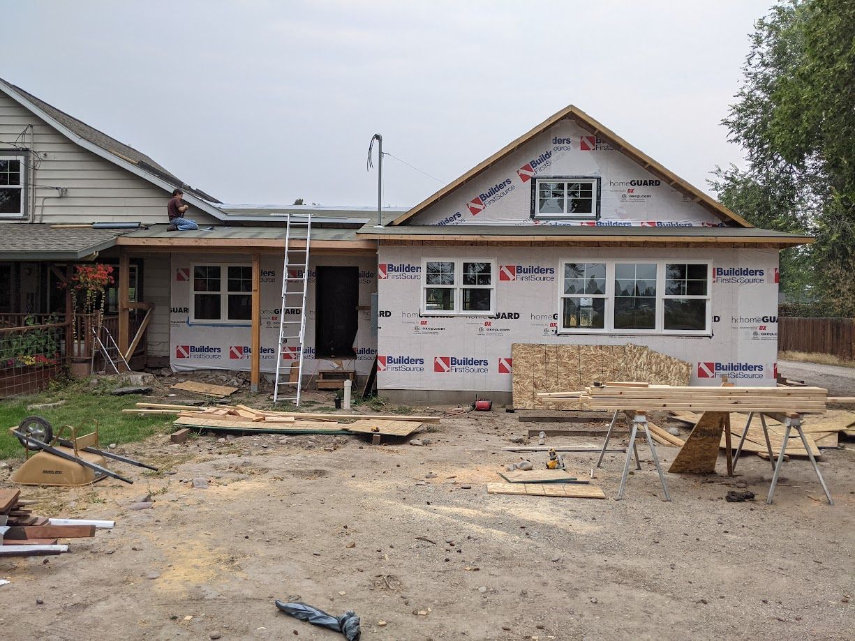 A house under construction with exposed exterior framing, white weather-resistant barriers, and windows installed.