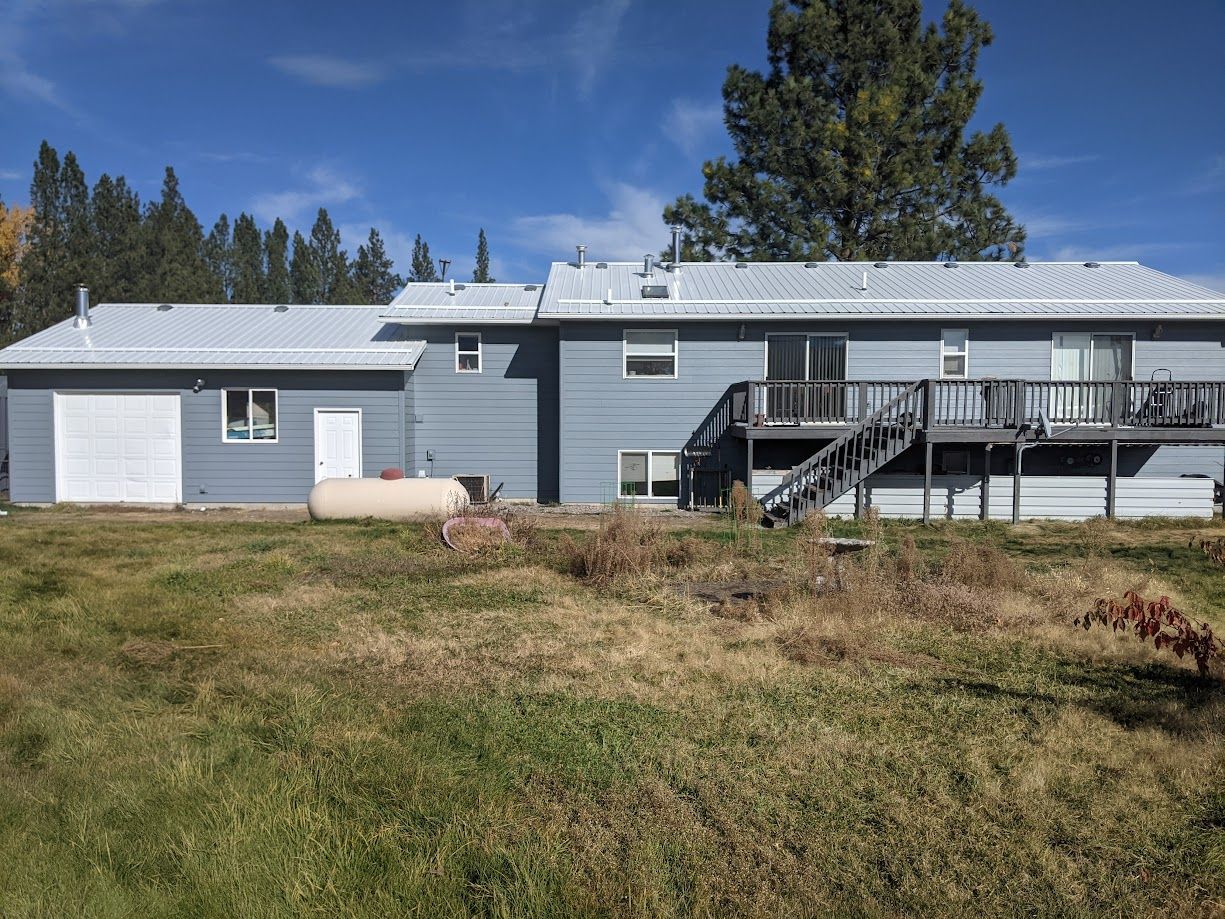 A light blue house with a white roof, a garage on the left, and a wooden deck with stairs in the back, set in a dry yard.