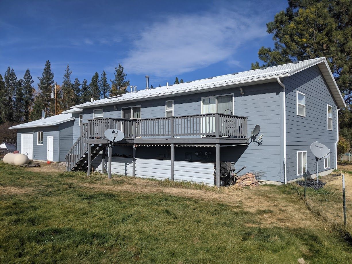 A grey, two-story house with a wooden deck and a white metal roof, situated on a grassy lot under a clear blue sky.
