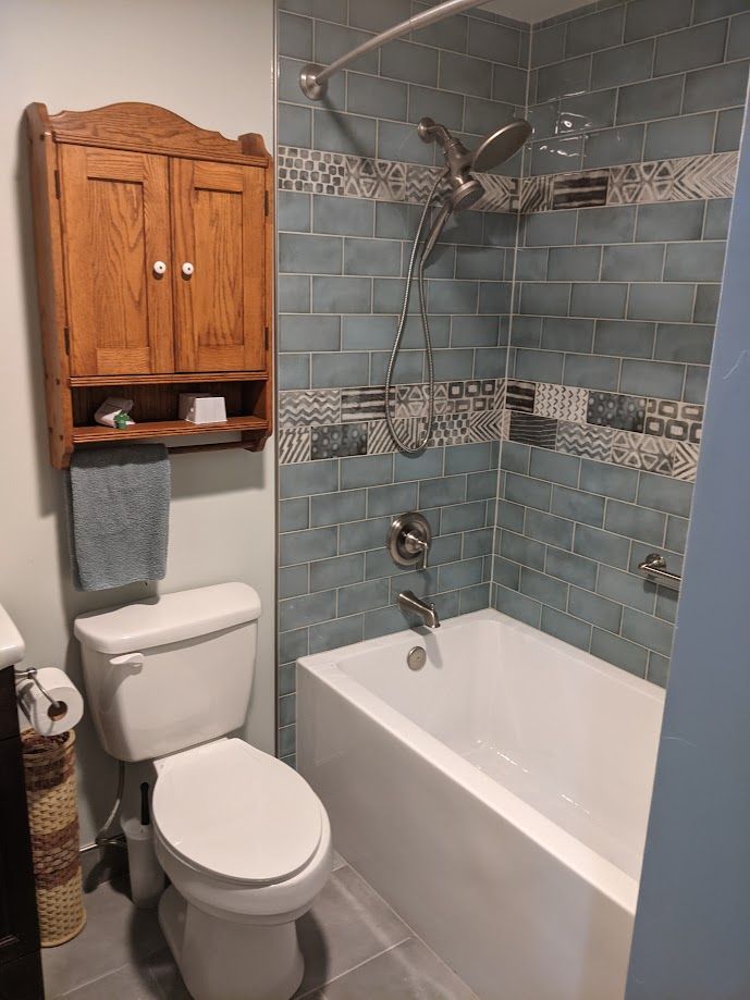 A bathroom with a white toilet, wooden wall cabinet, and a bathtub with blue subway tiles and a patterned accent strip.