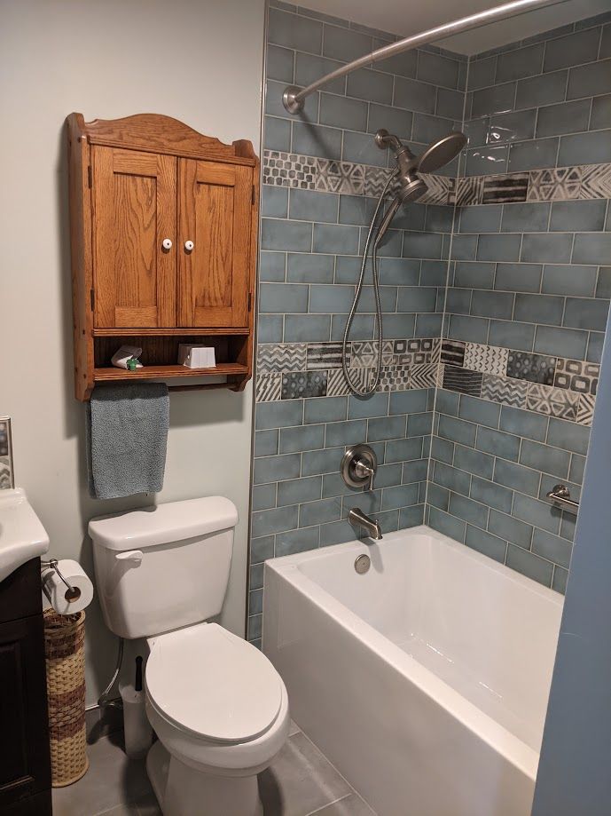 Small bathroom featuring a white toilet, wooden wall cabinet, and a bathtub with blue subway tiles and patterned accents.