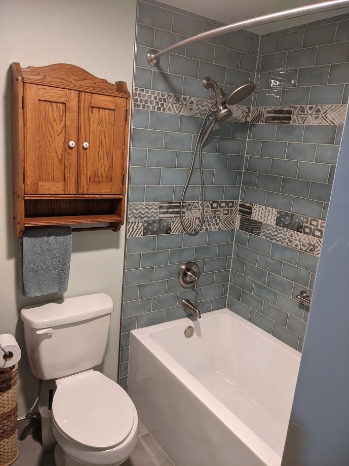 A bathroom with a white toilet, a wall-mounted wooden cabinet, and a bathtub surrounded by blue subway and patterned tiles.