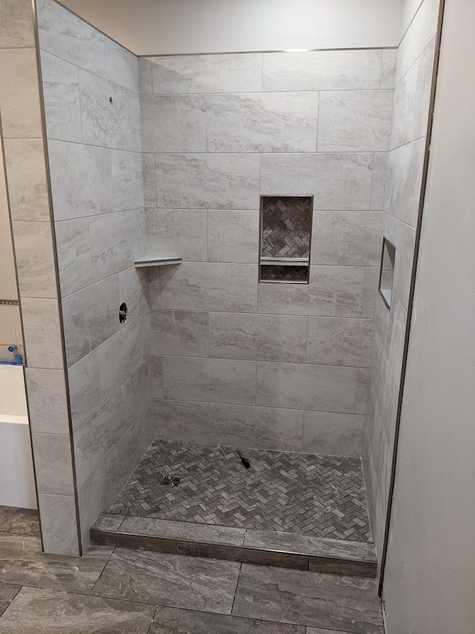 A walk-in shower with light grey tile walls, a patterned tile floor, a recessed wall niche, and a corner shelf.