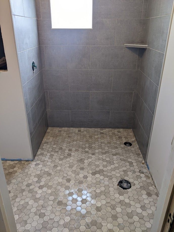 A walk-in shower with grey rectangular wall tiles, a light-colored hexagonal floor tile pattern, and two floor drains.
