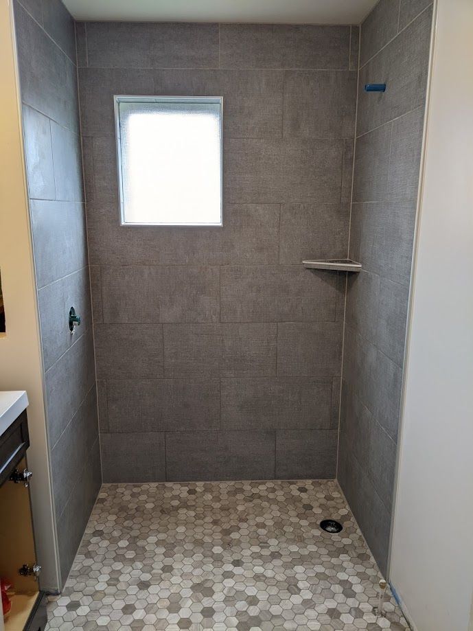 A walk-in shower with gray tiled walls, a small square window, a corner shelf, and a pebbled floor.