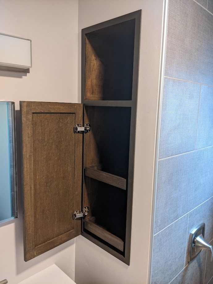 A recessed, dark wood cabinet with an open door, installed in a bathroom wall beside a tiled shower.